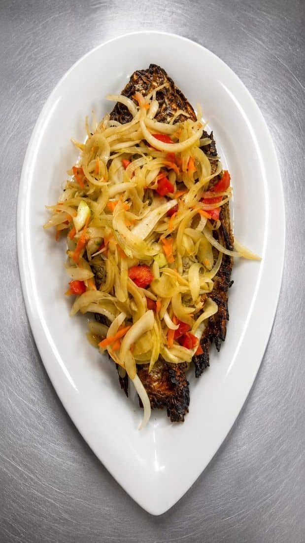Poisson braisé entier garni d'oignons et de piments, servi dans une assiette ovale blanche